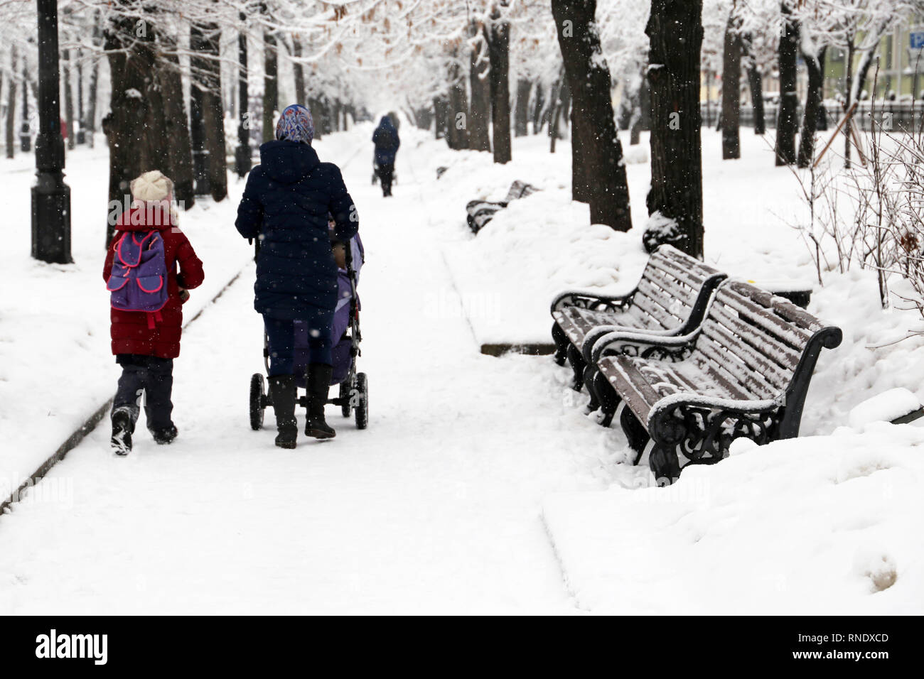 Mamá con un cochecito y una pequeña hija caminando en el Winter Park durante la nevada. Ir a la escuela, el clima de nieve, madre soltera Foto de stock