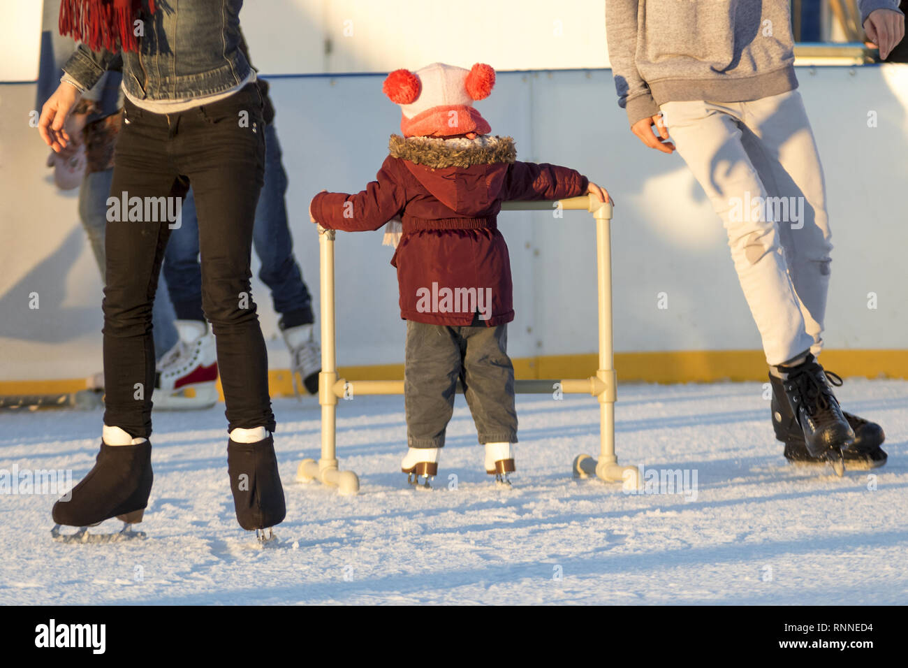 Niño pequeño aprende los primeros pasos sobre patines en la pista de patinaje sobre hielo. Una niña rodeada por adultos skaters mantiene el apoyo de estructura de skate en la pista de patinaje sobre hielo y l Foto de stock