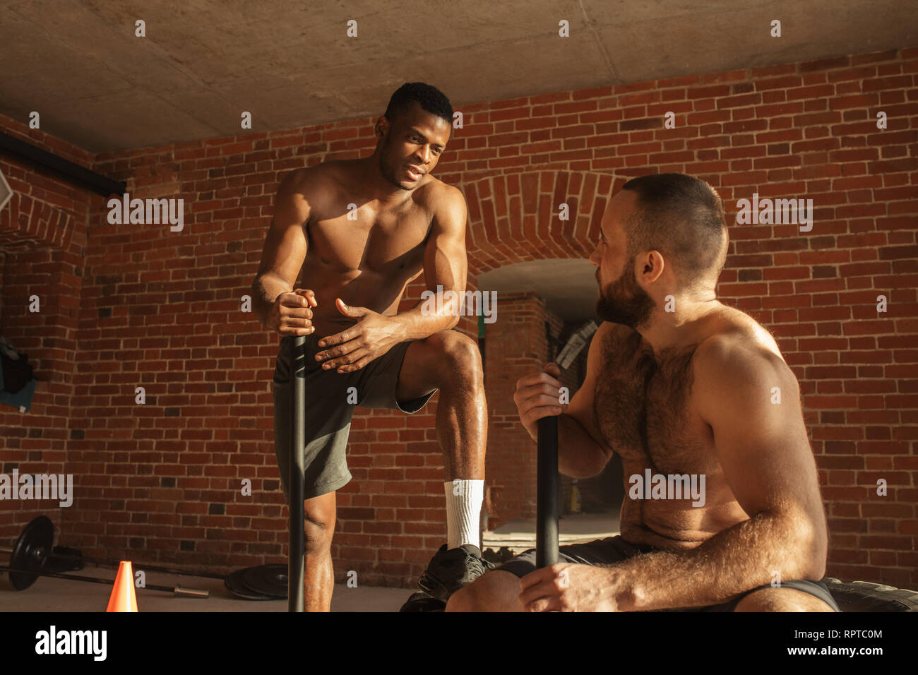 Dos mestizos los culturistas hablando durante un breve descanso en el  entrenamiento, los chicos están golpeando con un martillo neumático en  crossfit gimnasio con paredes de ladrillo y p Fotografía de stock -