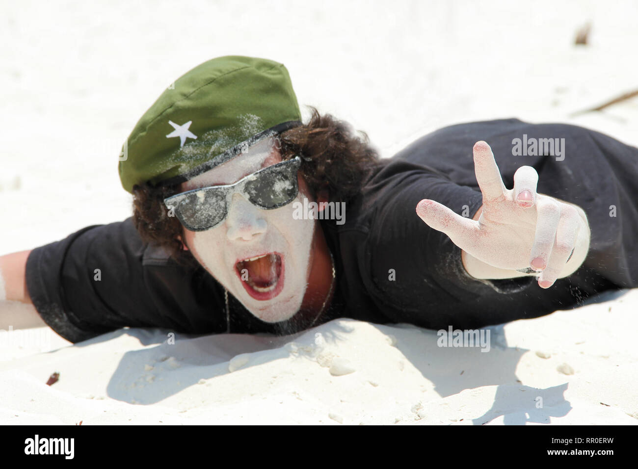 Un hombre joven en gafas de sol tumbado en una playa de arena blanca y travesuras, untando su rostro en la arena y gritando con alegría. El concepto de happ Foto de stock