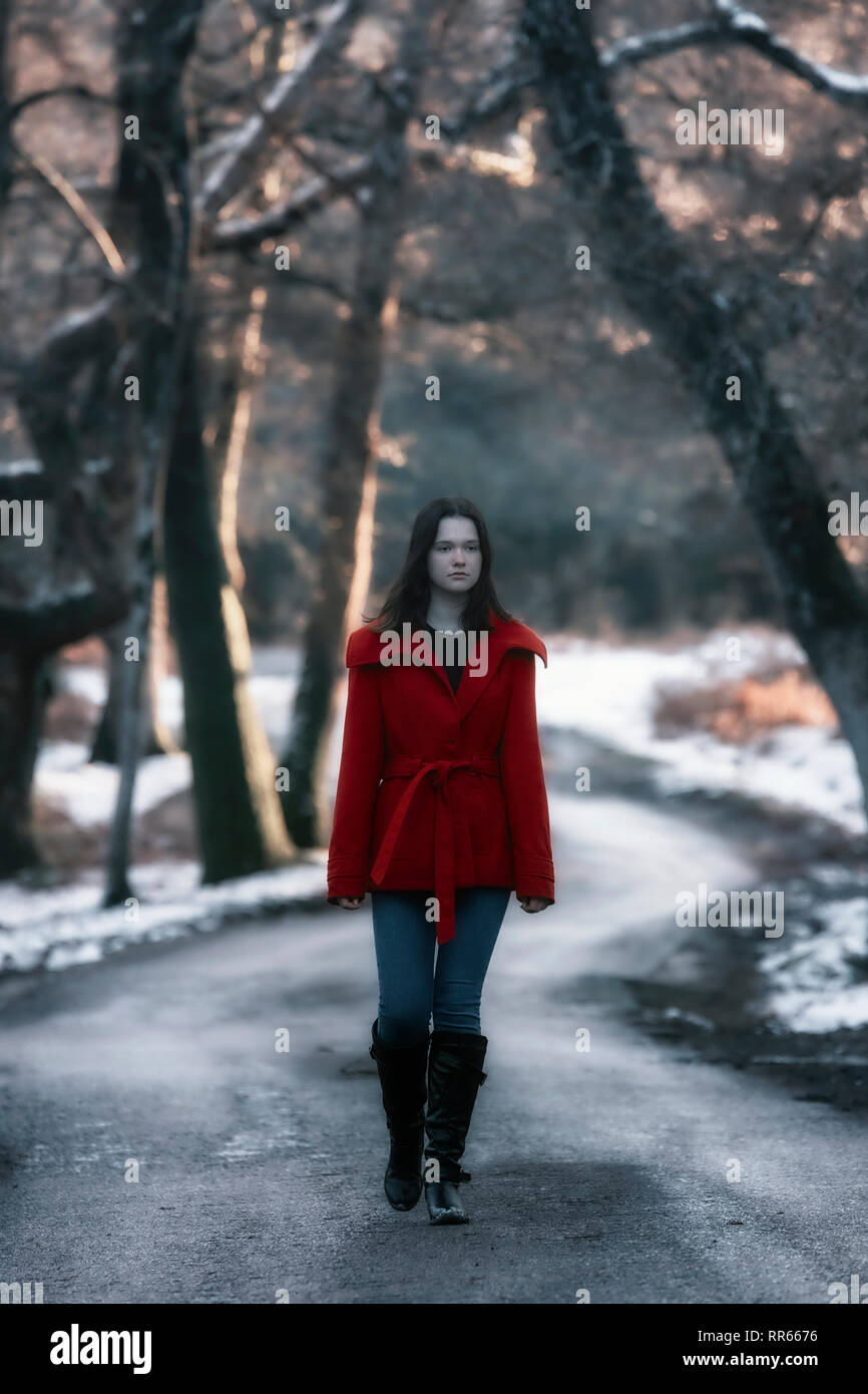 Una mujer joven con una chaqueta roja caminando por una calle pequeña en un bosque invernal con nieve Foto de stock