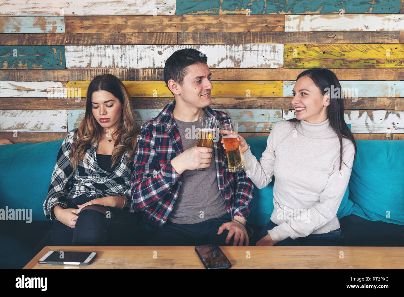 Feliz joven bebiendo cerveza con joven y socializar ignorando otros celoso triste mujer sentada junto a ellos en el rústico bar restaurante. Amor y j Foto de stock