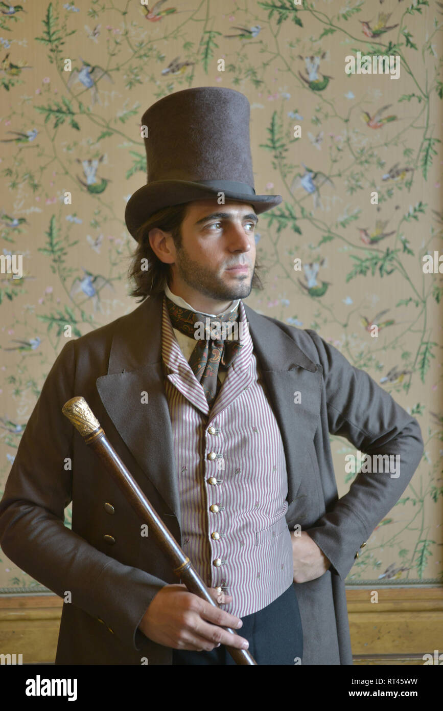 Un joven apuesto hombre Victoriano en un traje históricamente exacta está contra un telón de fondo de papel tapiz para un retrato- tiene un sombrero de y bastón Fotografía de stock -