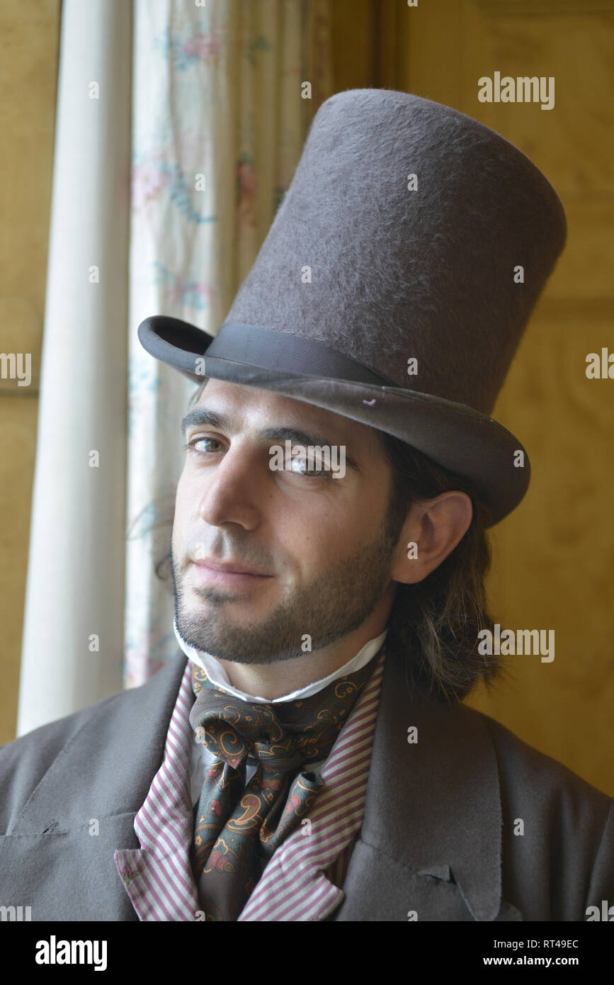 Un joven Victoriano en traje históricamente exacta está contra un telón de fondo de papel tapiz para un retrato- tiene un sombrero de copa y bastón Fotografía de stock -