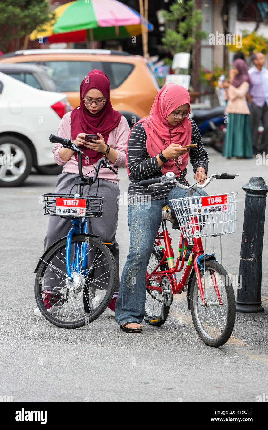 George Town, en Penang, Malasia. Jóvenes mujeres de Malasia sobre bicicletas comprobando sus teléfonos celulares. Foto de stock