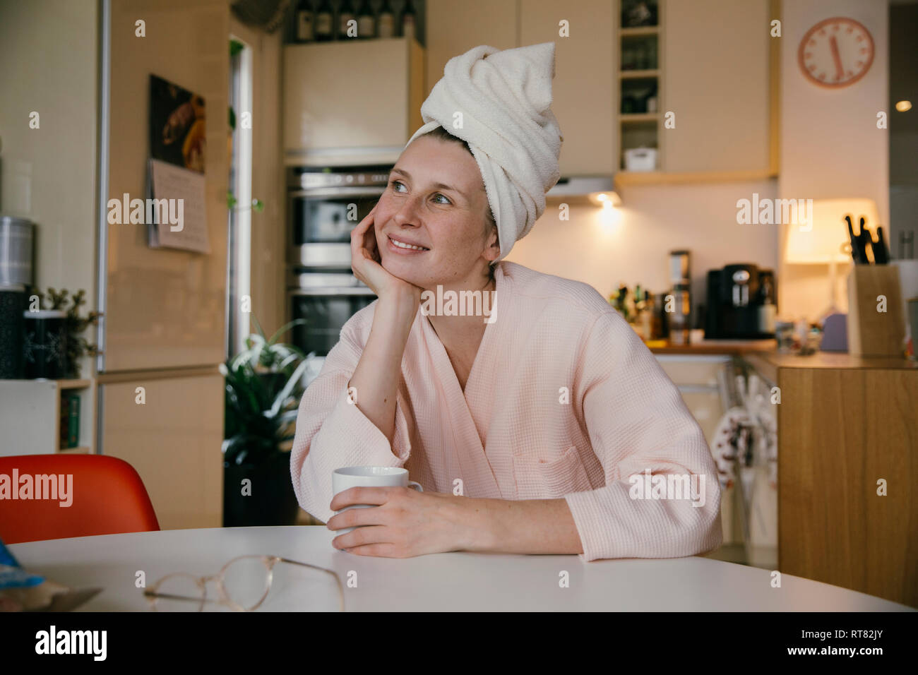 Retrato de mujer sonriente vistiendo una toalla turbante sentado con una taza de café en la mesa de la cocina Foto de stock