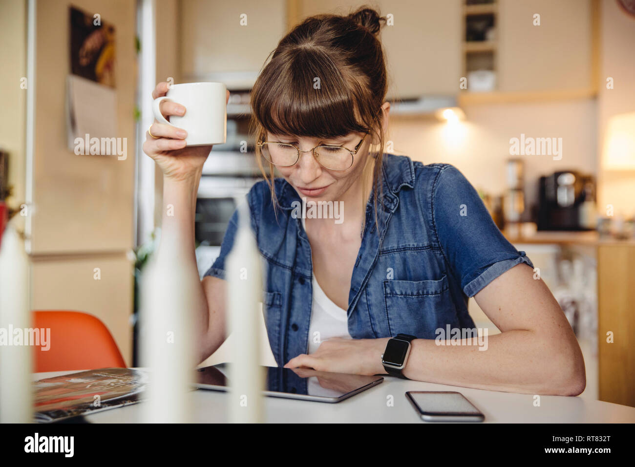 Mujer sentada con una taza de café en la mesa de la cocina utilizando tablet Foto de stock