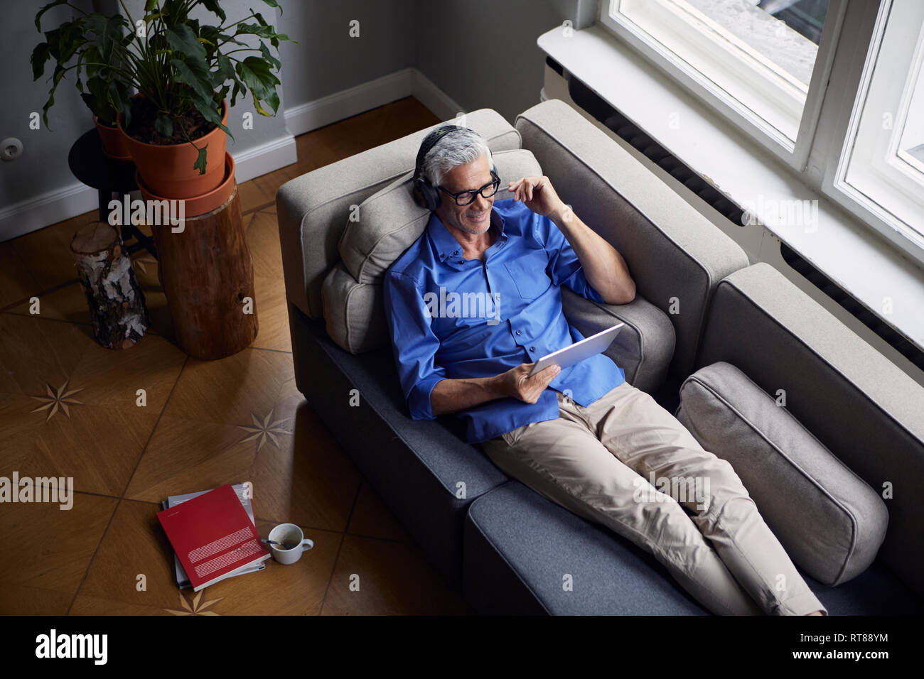 Sonriente hombre maduro con tablet y auriculares relajándose en sofá en casa Foto de stock