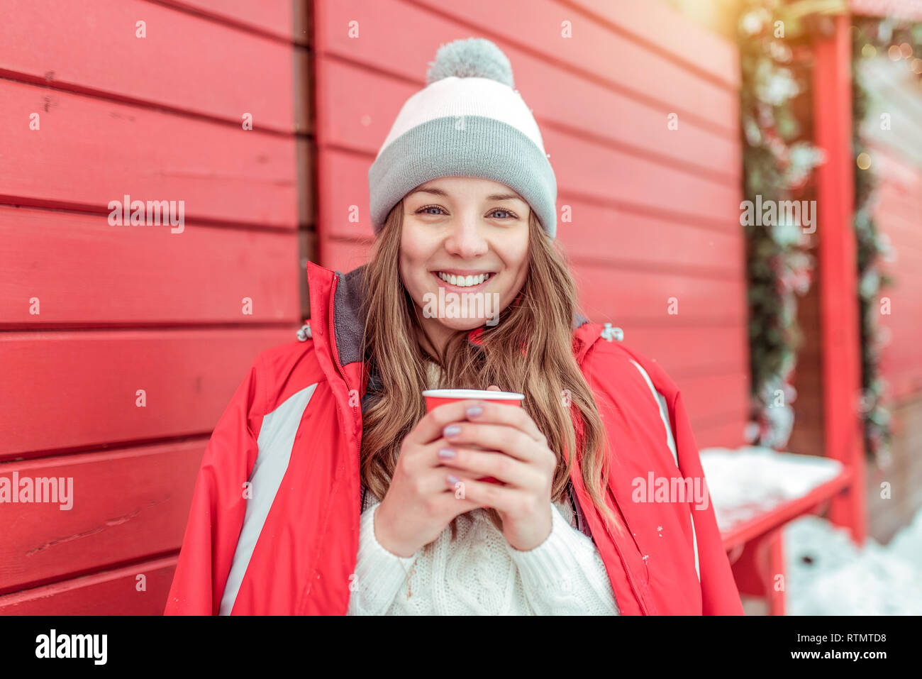 Bella mujer en invierno en una chaqueta sobre un fondo de color rojo con contraventanas de madera. Sosteniendo una taza de café, té. Feliz sonriendo y riendo. Holiday Resort Foto de stock