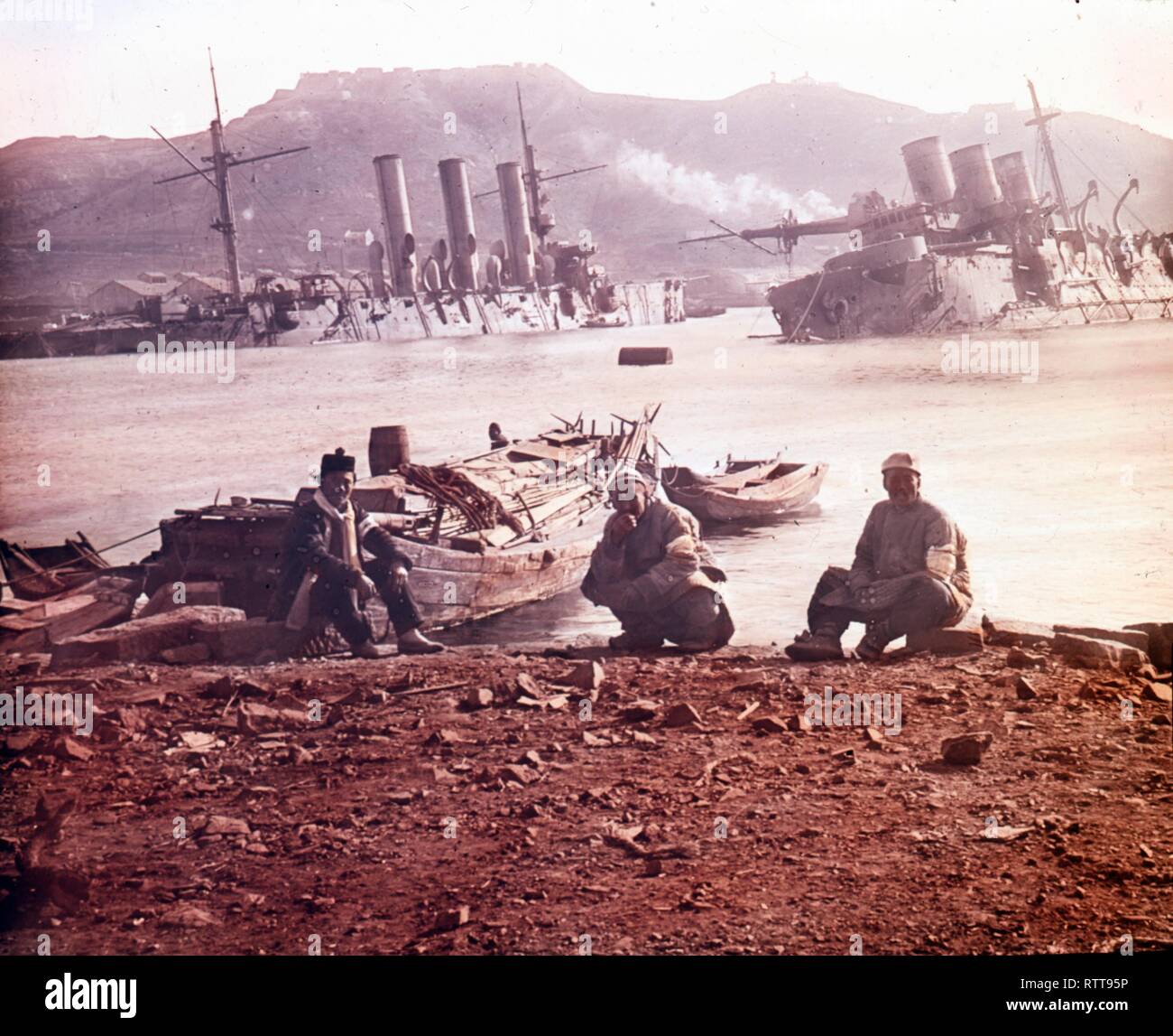 Foto coloreada de tres soldados japoneses sentados en una playa junto al puerto de Port Arthur cerca de un buque de guerra ruso hundido durante la Guerra Ruso-Japonesa, Port Arthur, China, 1905. (Foto de Burton Holmes) Foto de stock