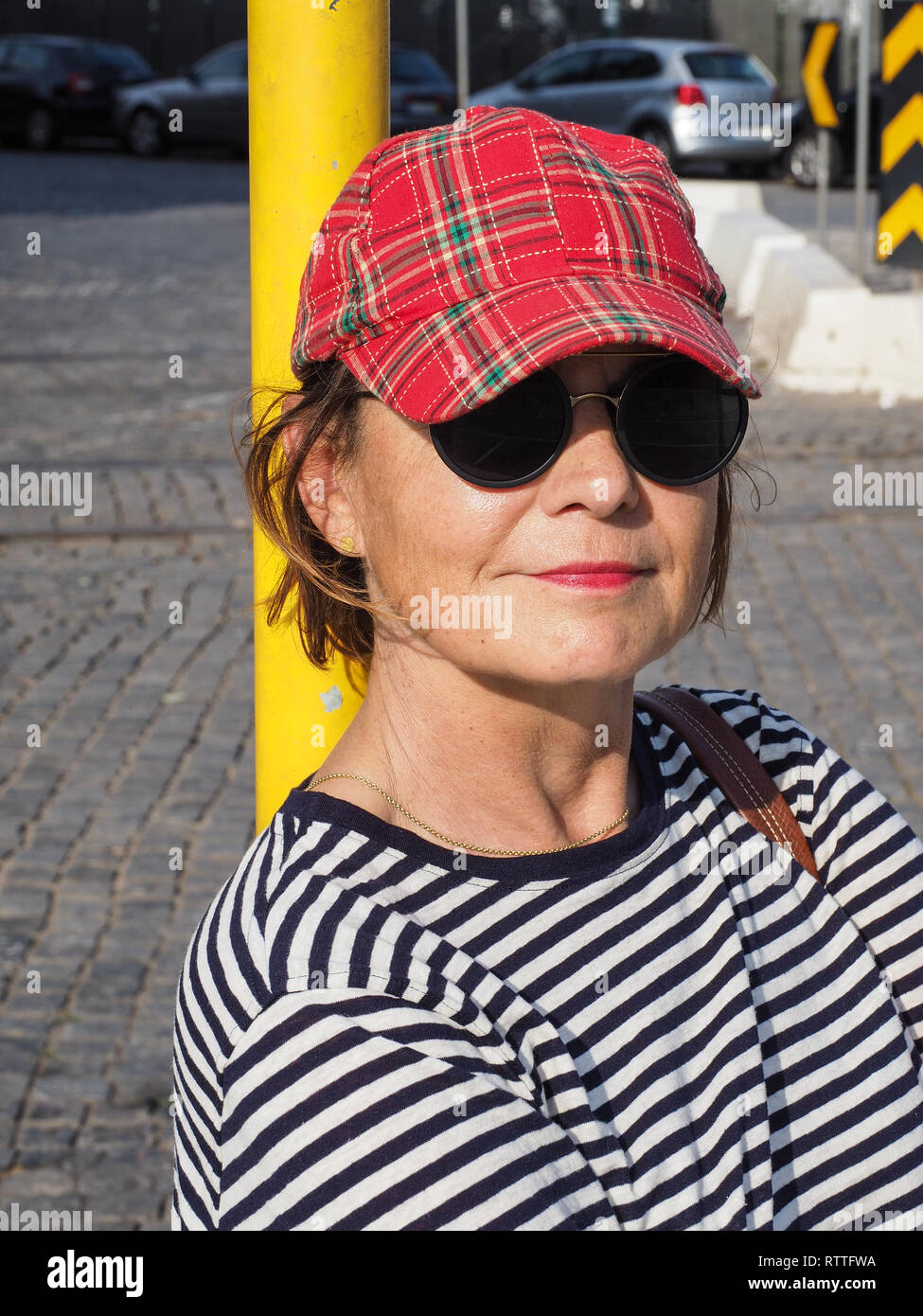 Mujer feliz con gafas de sol, una gorra de cuadros y una camisa a rayas, apoyado contra un polo amarillo mirando a la cámara con una amplia y cálida sonrisa Foto de stock