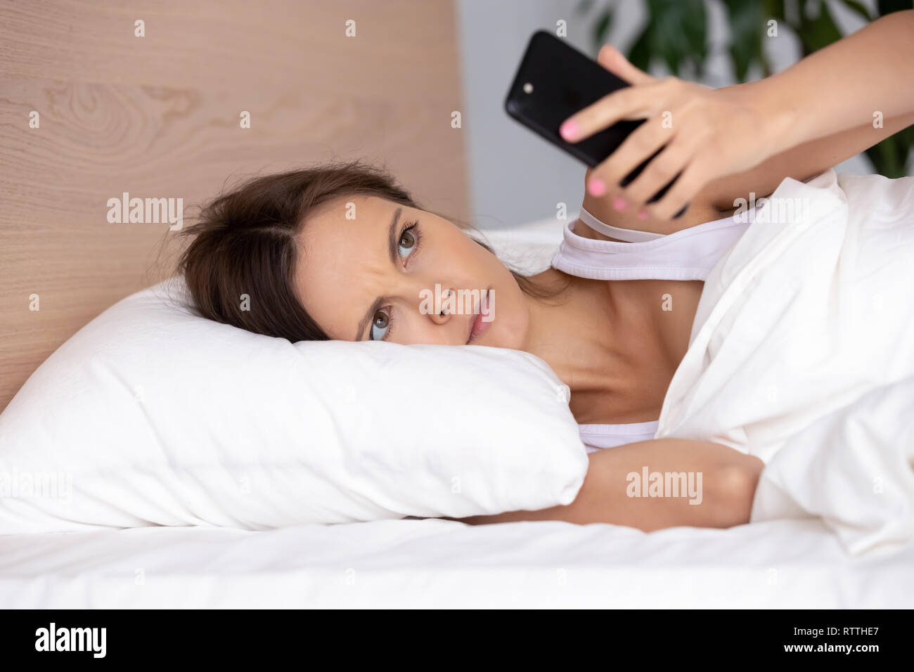 Malestar joven utilizando el teléfono en la cama, mirando a la pantalla, insomnio Foto de stock