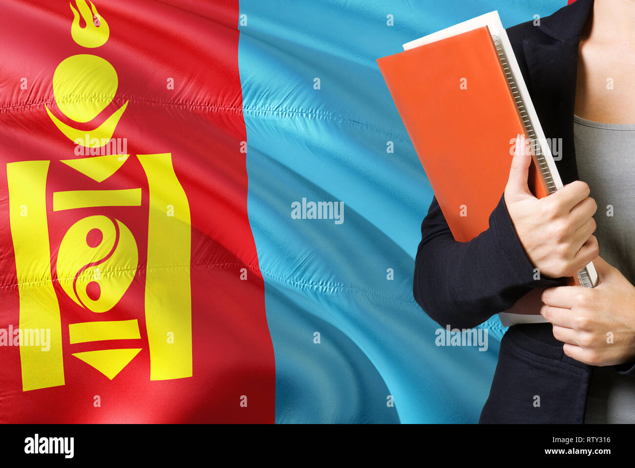 Aprender mongol concepto. Mujer joven de pie con la bandera de Mongolia en el fondo. Profesor sosteniendo libros, naranja cubierta de libro en blanco. Foto de stock