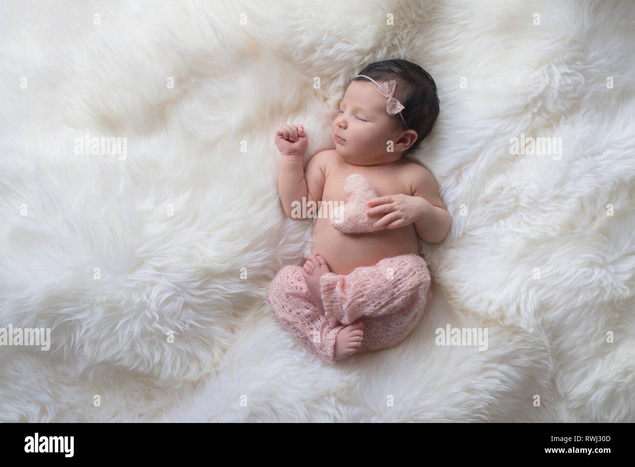 Dormir, semana de edad bebé vestida de luz rosa, tejida de pantalones y sosteniendo un minúsculo, almohada en forma de corazón. Filmado en el estudio sobre un blanco sheepski Foto de stock