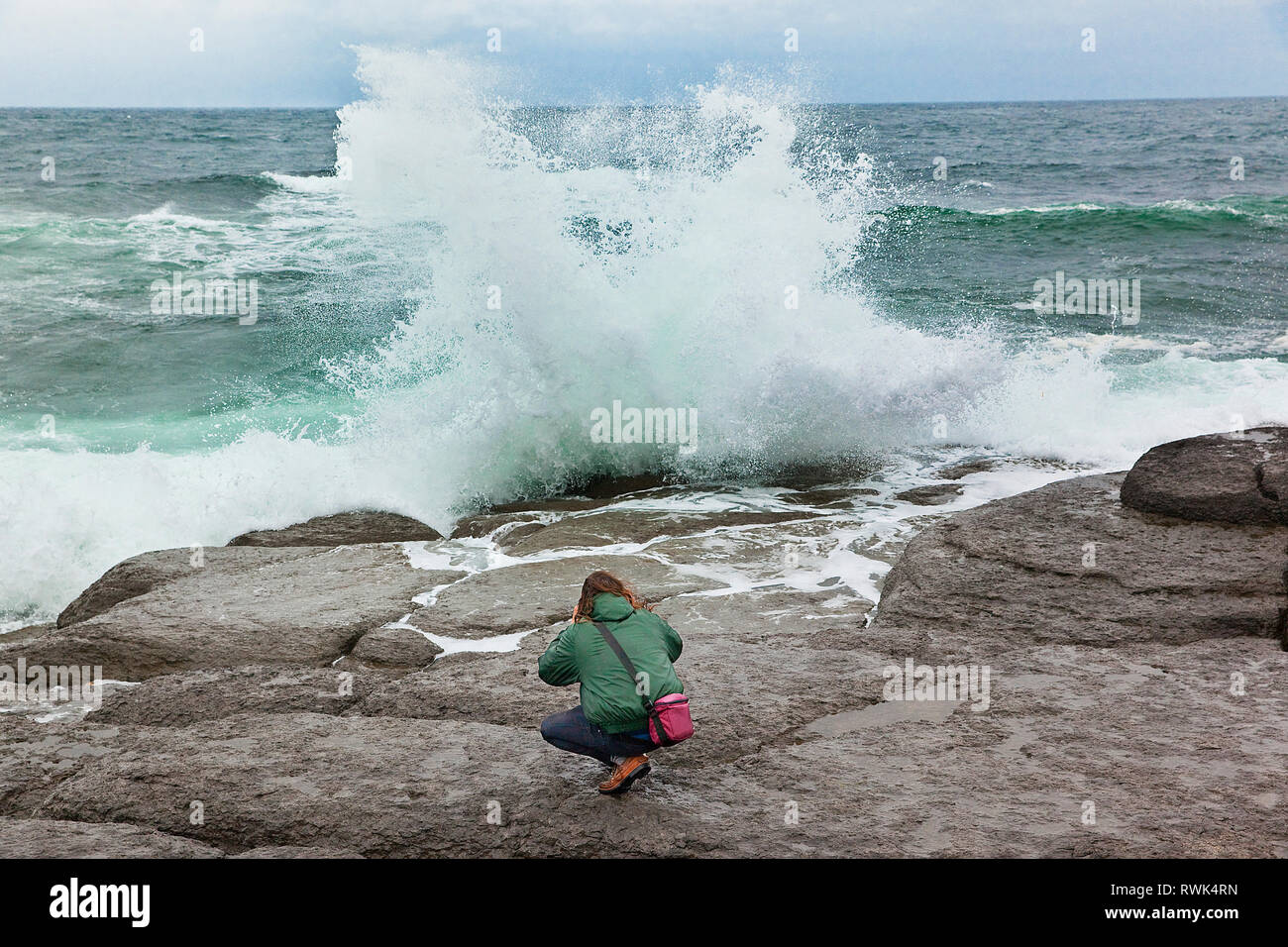 Joven en cuclillas sobre una roca saliente, en un intento de fotografiar el romper de las olas de cerca. Port au Choix National Historic Site, Port au Choix, Newfoundland, Canadá Foto de stock