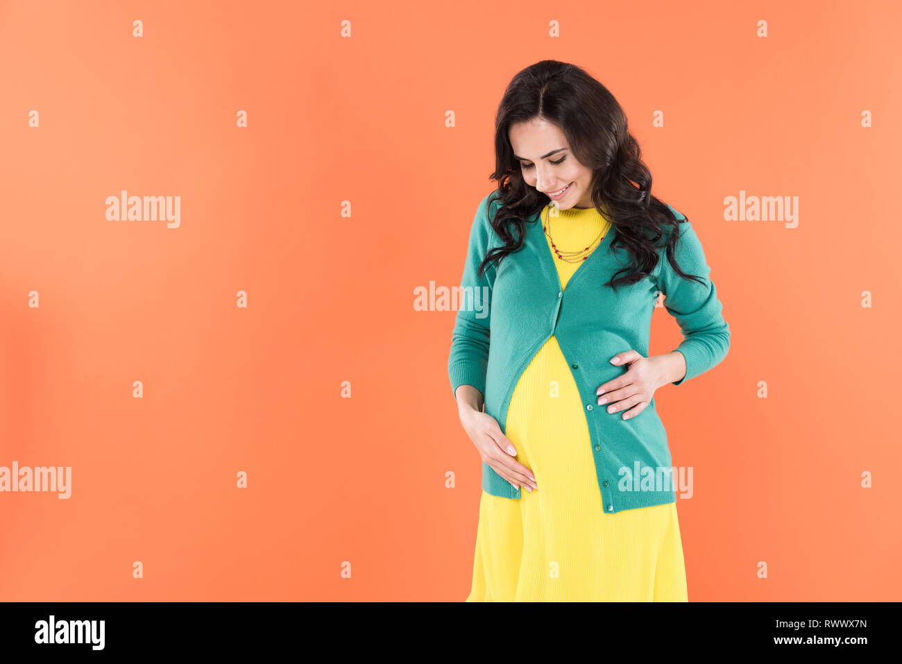 Dichosa la mujer embarazada en ropa brillante tocando el vientre aislado en naranja Foto de stock