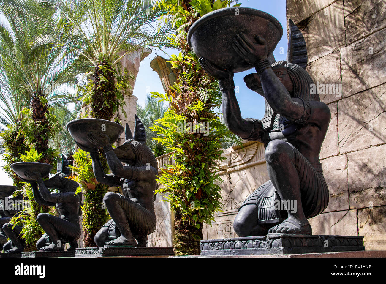 Callejón con grandes estatuas de piedra idéntica del dios egipcio Anubis, quien se halla en una rodilla y sostiene un cuenco redondo en sus manos. Las palmeras crecen entre th Foto de stock