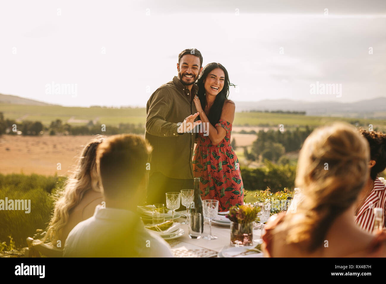 Pareja amorosa en una fiesta anunciando su matrimonio con sus amigos. Hombre y mujer sonriente haciendo un anuncio durante una cena al aire libre. Foto de stock