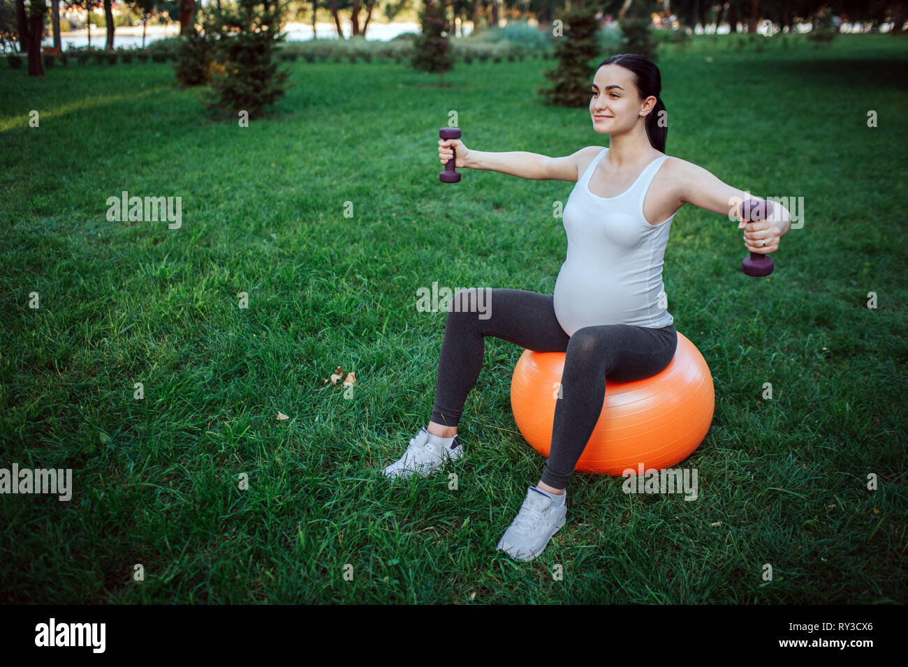 Bonita alegre joven embarazada sentarse sobre la bola de fitness naranja en parl. Ella ejercicio con pesas. Foto de stock