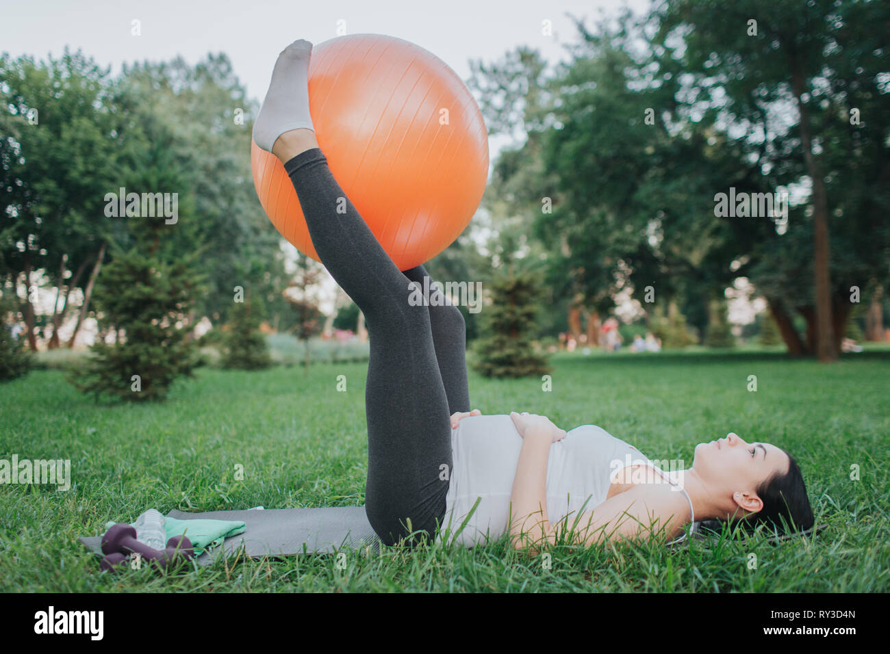 Joven Embarazada exerising concentrada sobre yoga mate en el parque. Que ella sostenga naranja grande pelota fitness entre las piernas. Modelo mantenga las manos sobre el vientre. Foto de stock