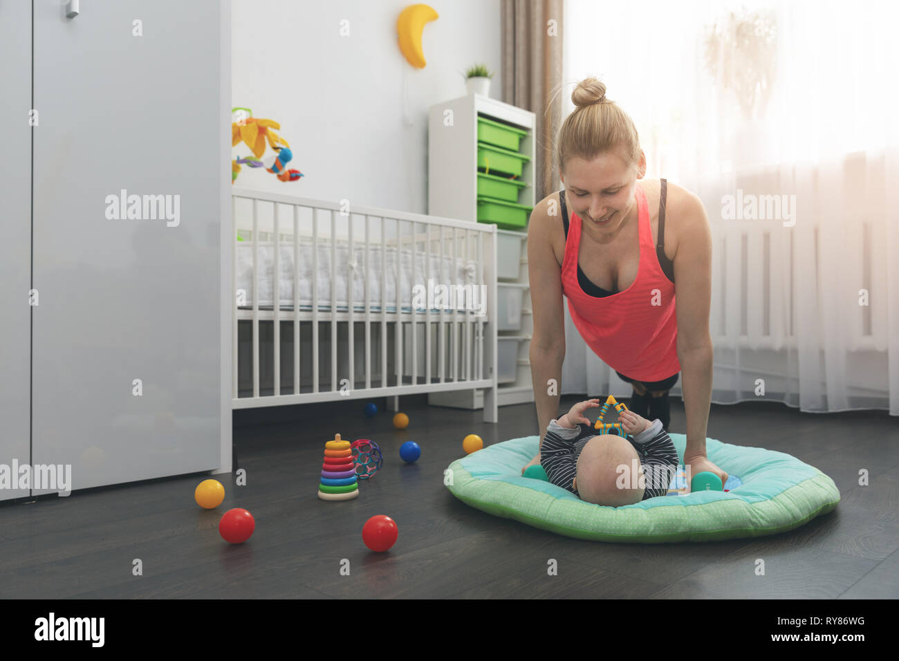 Mujer joven haciendo flexiones en casa mientras juega con su pequeño bebé Foto de stock