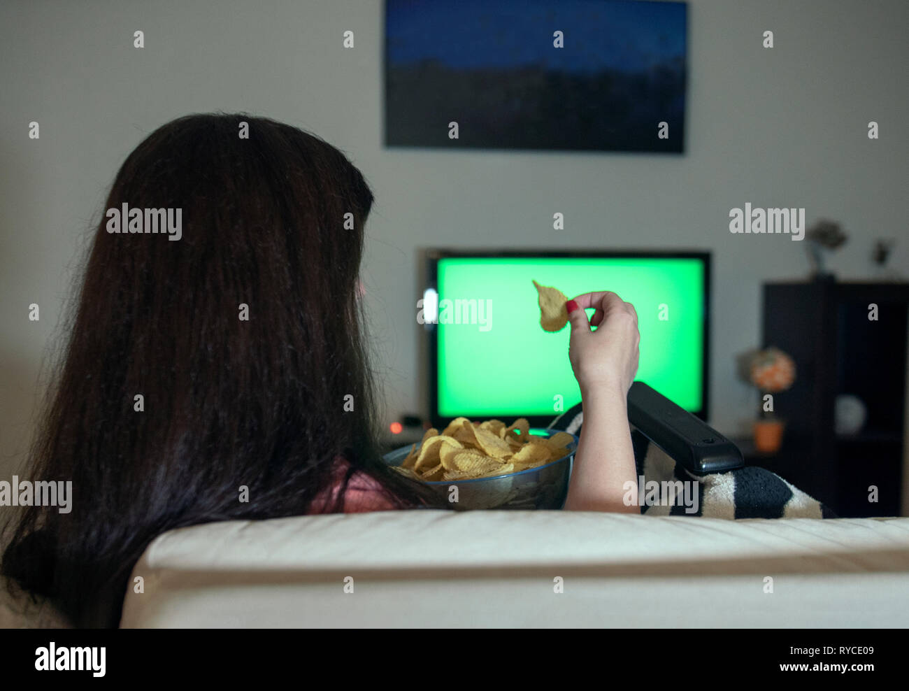 Mujer morena sentada en su casa relajante noche comiendo papas fritas y viendo televisión, chromakey Foto de stock