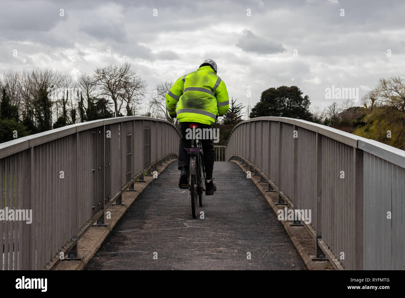 El hombre monta su bicicleta sobre un puente después del trabajo llevaba una chaqueta de alta visibilidad Foto de stock