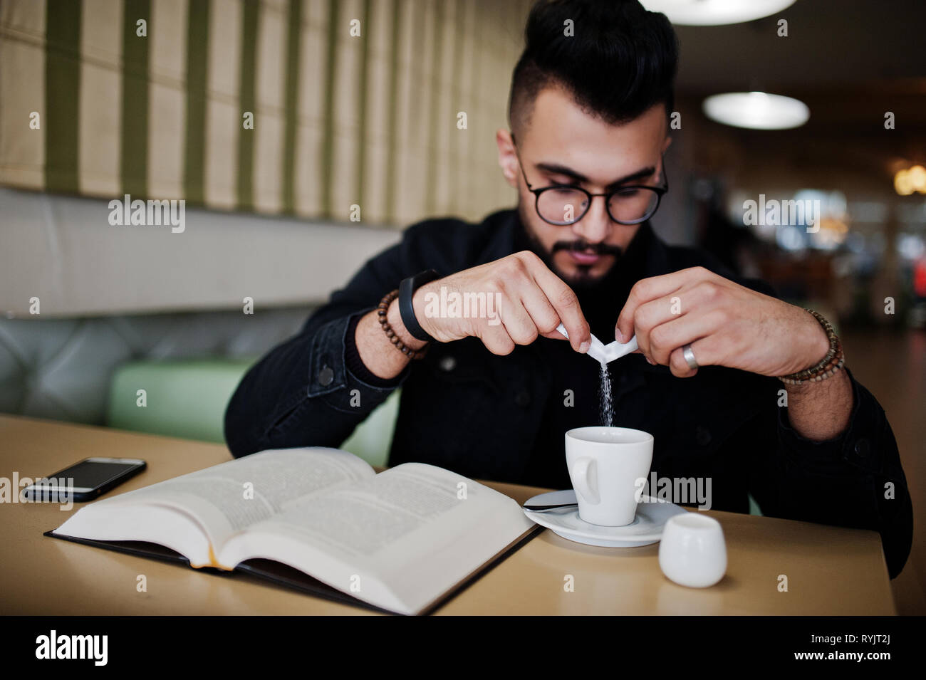 Formación Parásito Guardería Hombre árabe desgaste en negro chaqueta jeans y anteojos sentado en la  cafetería, leer libros y beber café. Elegante y moderno modelo arábigo guy.  Vierta el azúcar Fotografía de stock - Alamy