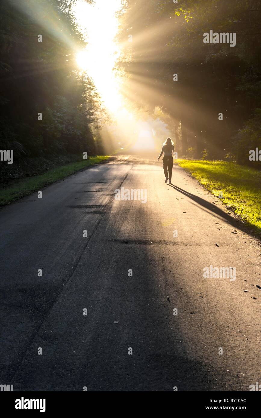 Mujer joven caminando por una calle, la luz del sol brilla a través de los árboles, Oregon Coast Highway, Oregón, EE.UU. Foto de stock