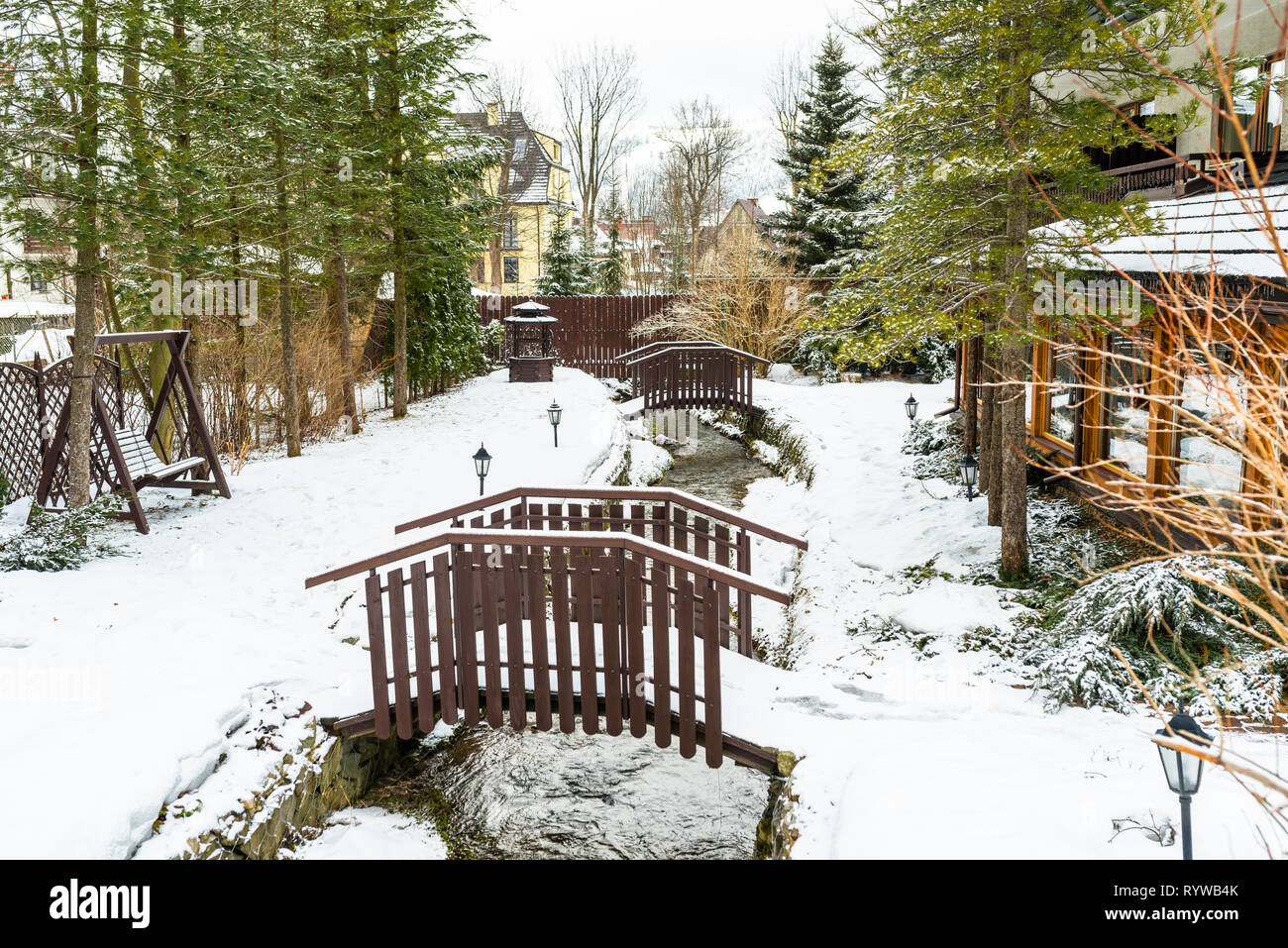 Paisaje invernal en la ciudad, la nieve cubrió el terreno visible dos pequeños puentes a lo largo de un arroyo de montaña verde y árboles coníferos. Foto de stock