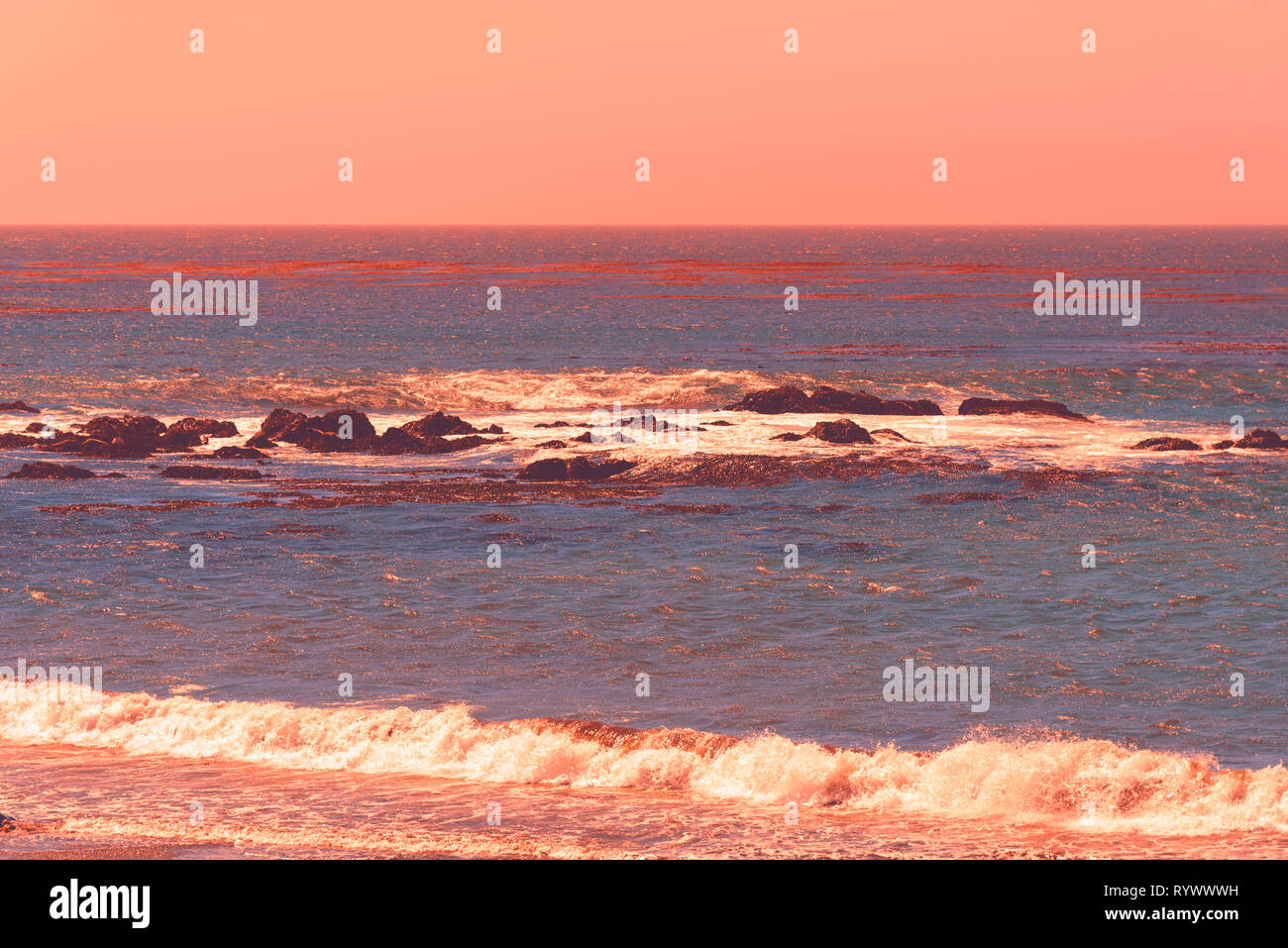 Océano Azul con olas rompiendo sobre las rocas, bajo cielos naranja y rosa pastel. Foto de stock