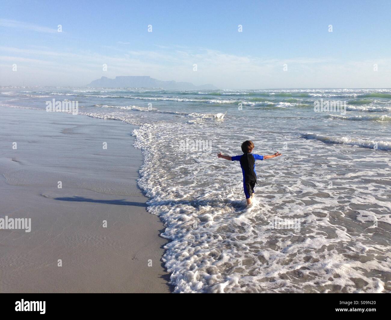 Joven jugando en el mar cerca de Melkbosstrand, Cape Town, Sudáfrica, con la montaña de la Mesa visibles en el fondo. Foto de stock