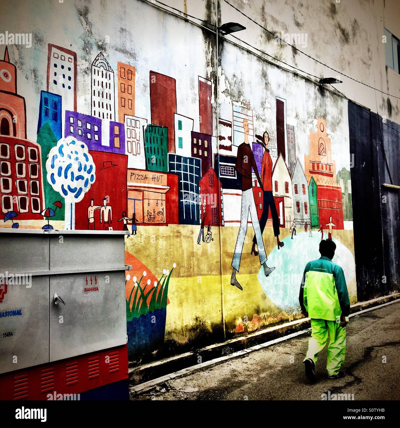 Hombre caminando por un muro decorado con murales pintados y fotografías y un panel eléctrico de Tenanga Nasional, Malasia Foto de stock