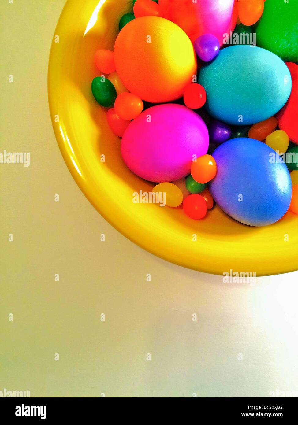 Un tazón de brillantes huevos teñidos y jelly beans en un tazón visto desde arriba sobre fondo blanco. Foto de stock