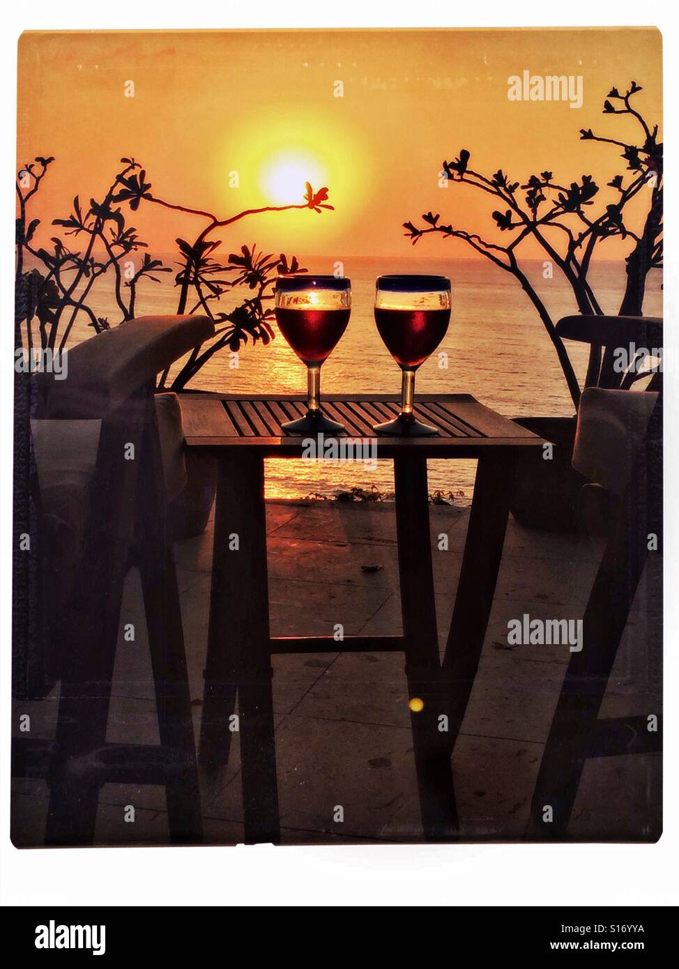Montura azul mexicano dos vasos de vino se rellenan con vino tinto sobre una tabla de madera de teca entre dos sillas en una terraza con vistas a la puesta de sol sobre el Océano Pacífico en Nayarit, México. Foto de stock