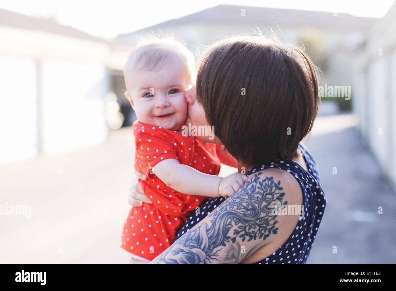 Hip joven mamá con el pelo corto y los tatuajes celebración niña hija besarla en la mejilla Foto de stock