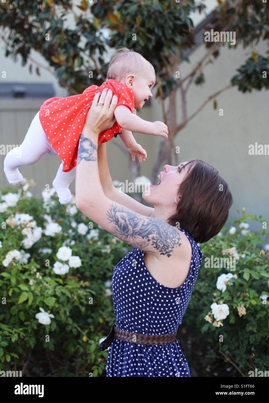 Hip joven mamá con el pelo corto y los tatuajes celebración niña hija en el aire sonriendo Foto de stock