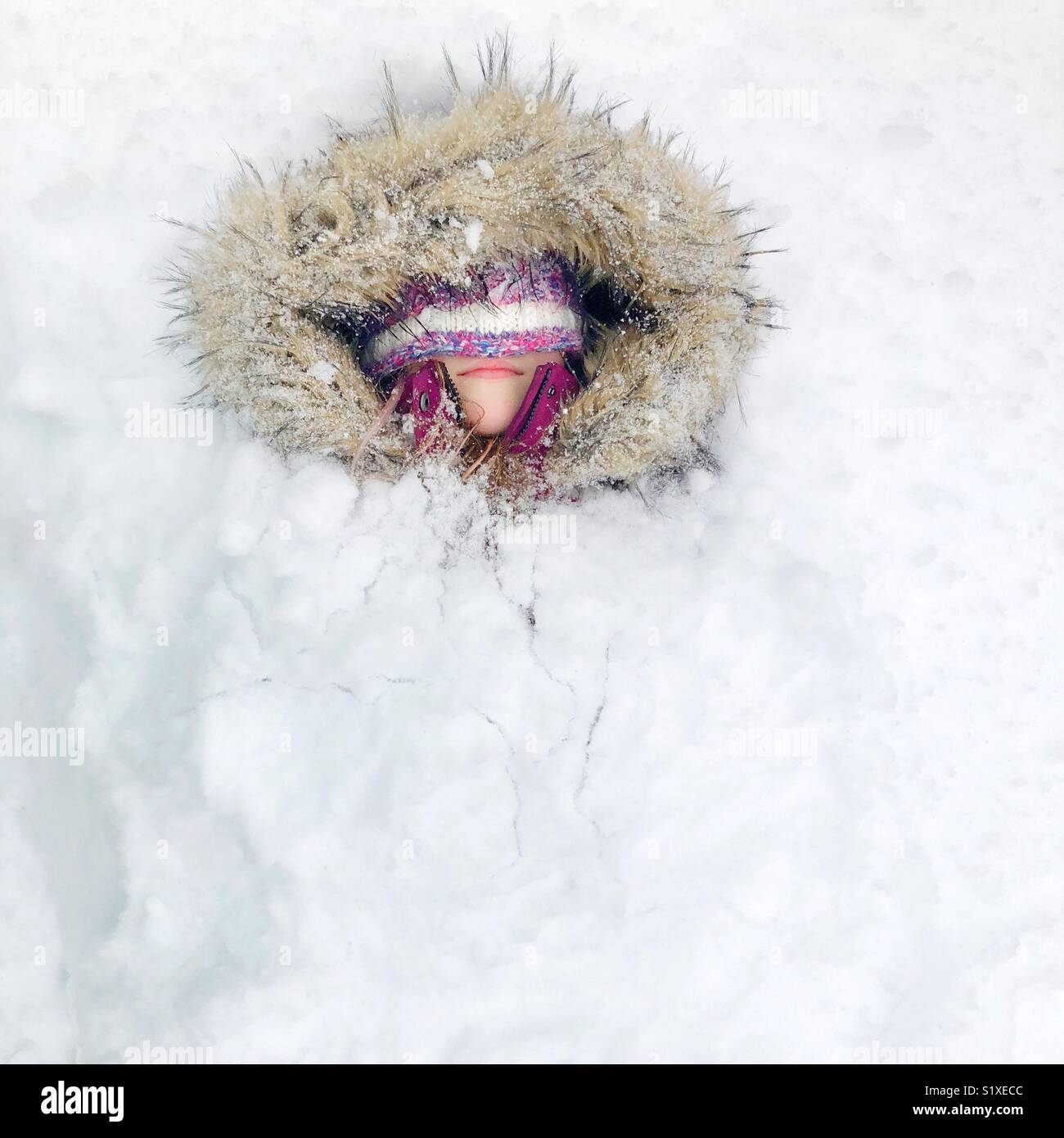 Chica con piel falsa cubierta y hat cubriendo ojos enterrados en la nieve mostrando sólo cabeza Foto de stock