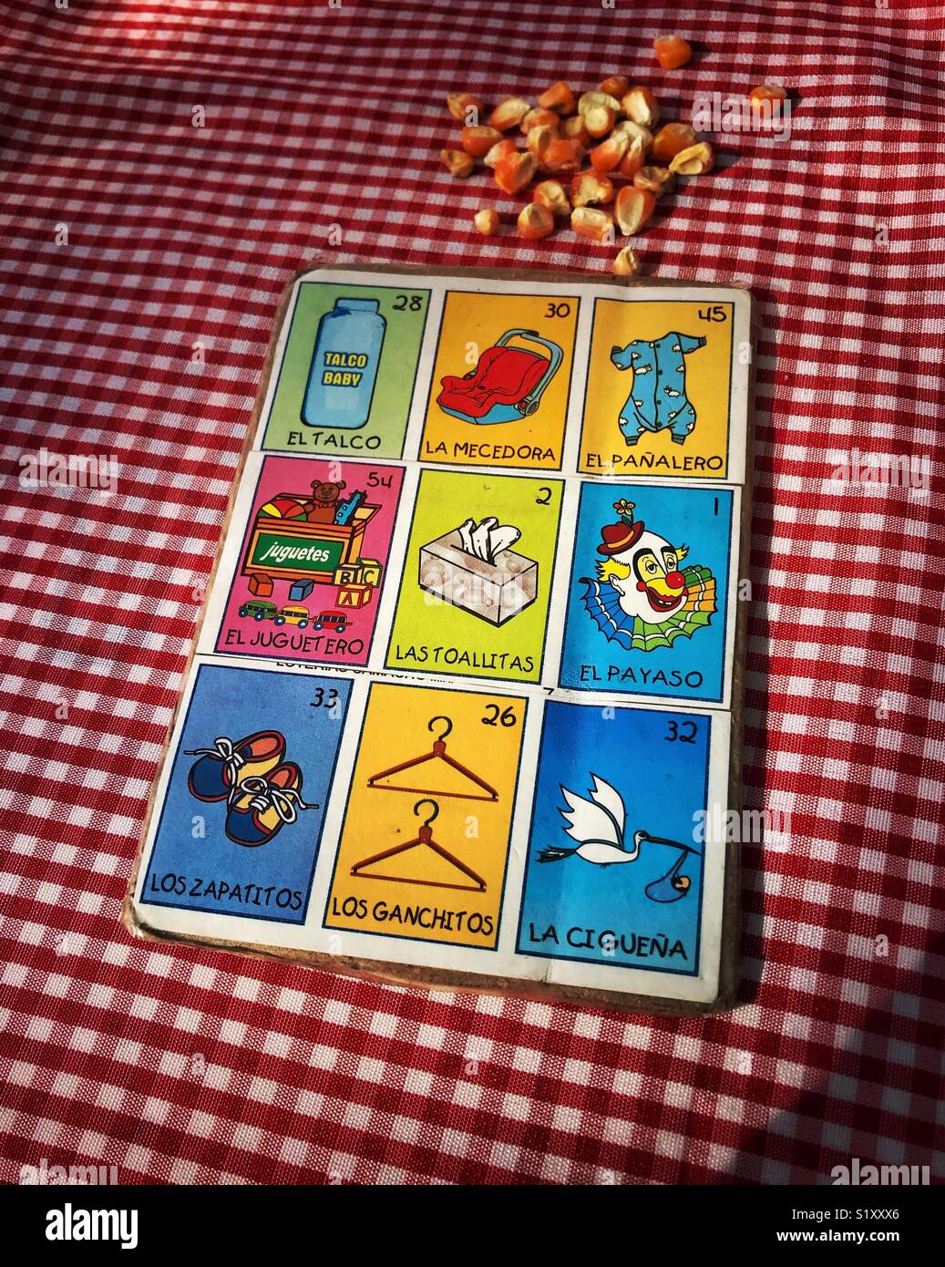Un mexicano baby shower Loteria bingo card y granos de maíz para los marcadores están establecidos en un mantel de cuadros rojo y blanco antes de que el juego comienza en un baby shower en la luz de la tarde. Foto de stock