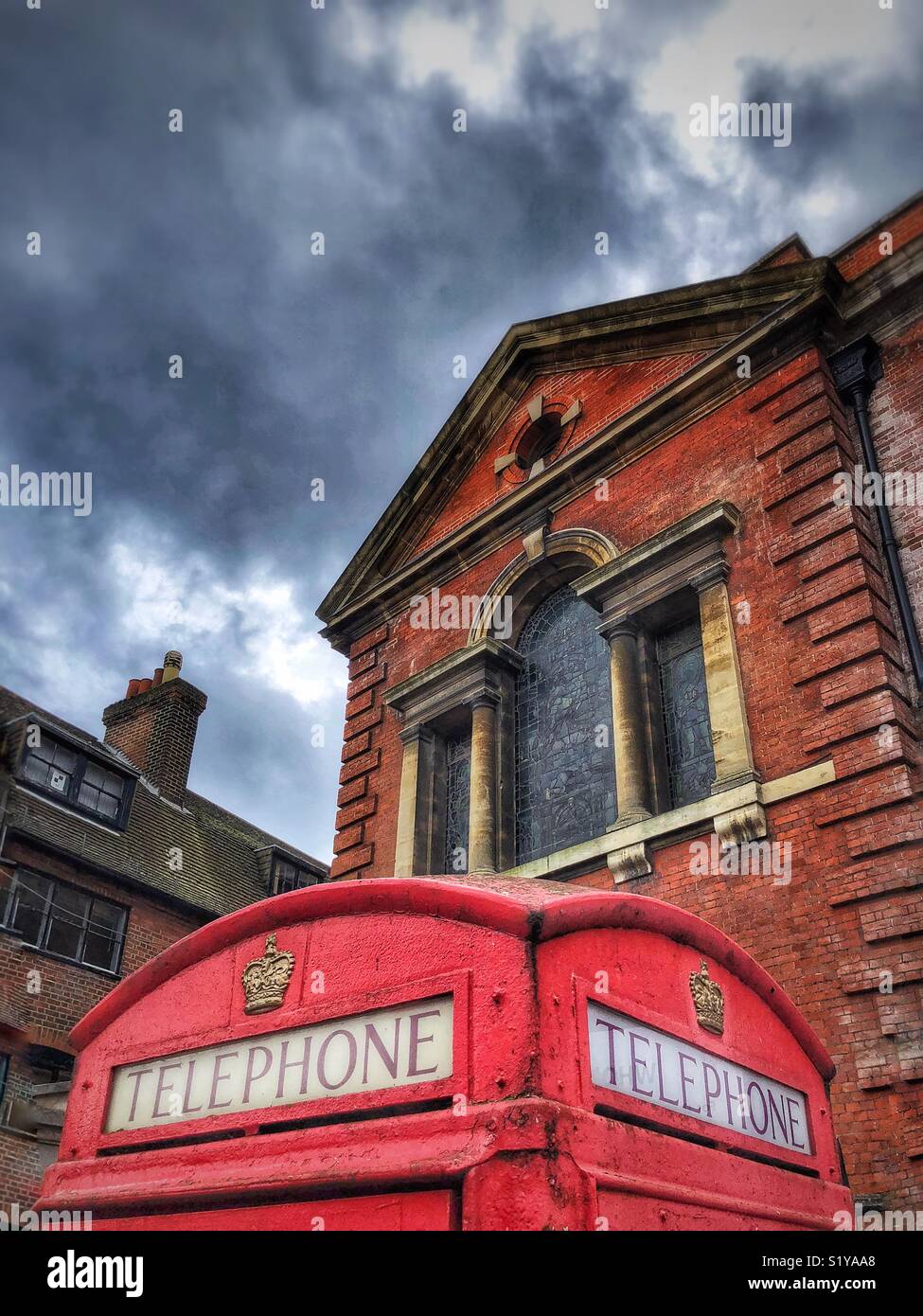 Cuadro teléfono rojo tradicional inglés contra el techo de un edificio tradicional y cielo tormentoso Foto de stock