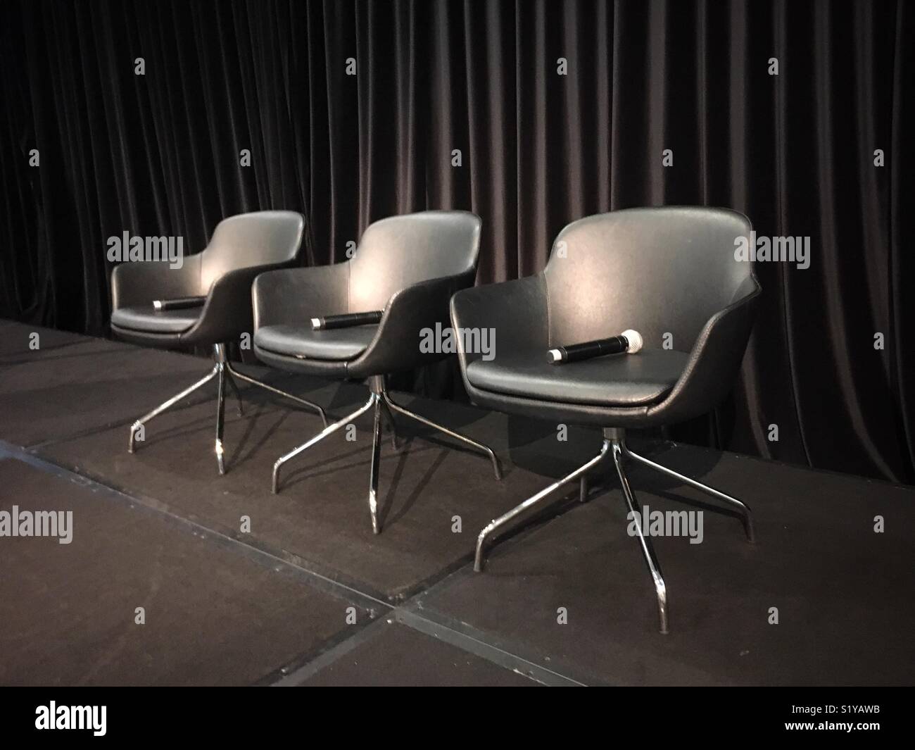 Tres sillas con micrófonos contra una cortina negra. Foto de stock