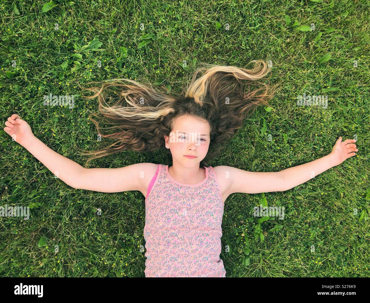 11 años de edad, niña recostada en la hierba con los brazos abiertos y grave expresión distante en la cara Foto de stock