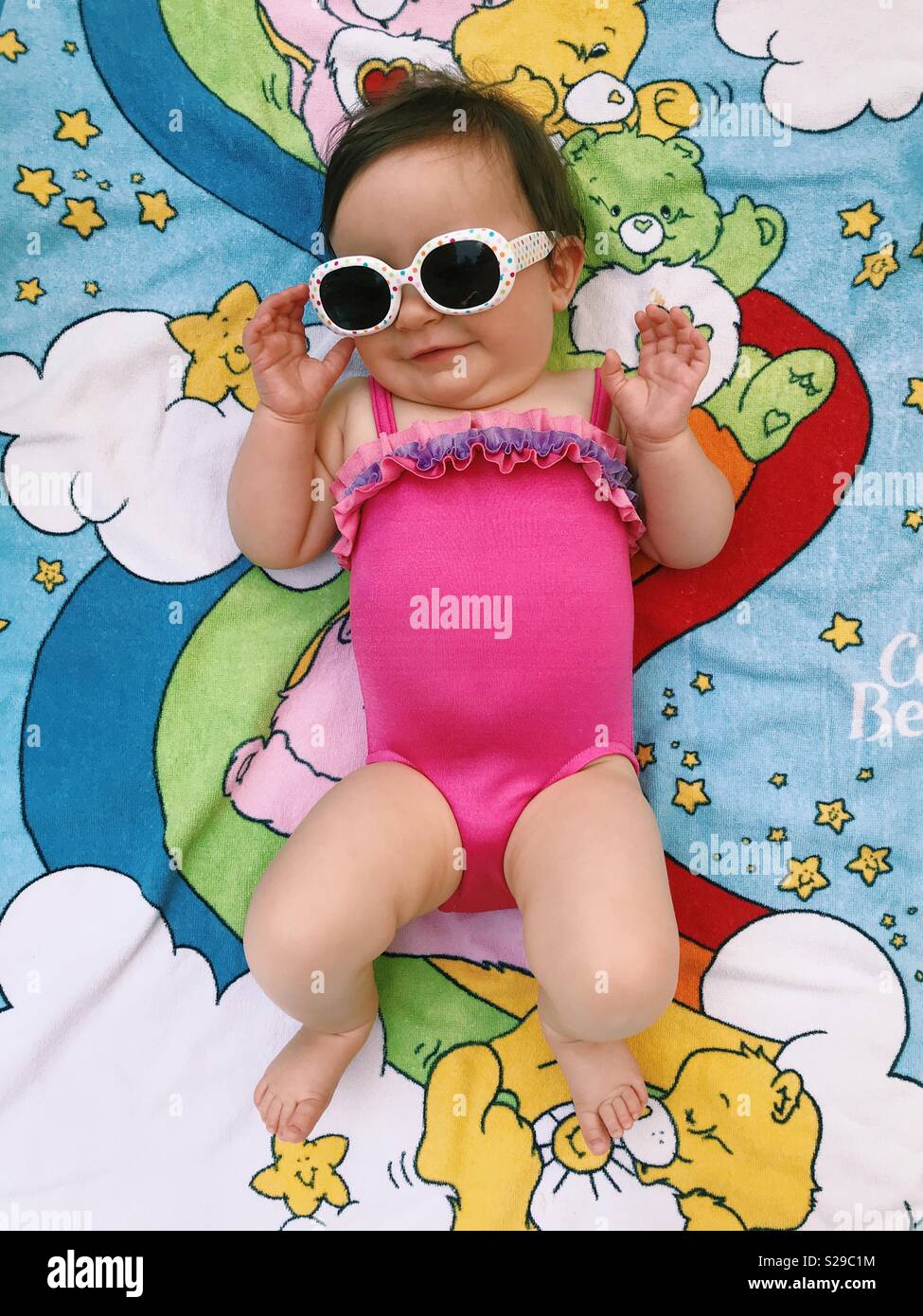 Un lindo bebé niña en un traje de baño rosa, con gafas de sol y tumbado sobre un cuidado Lleva una toalla de playa. Foto de stock