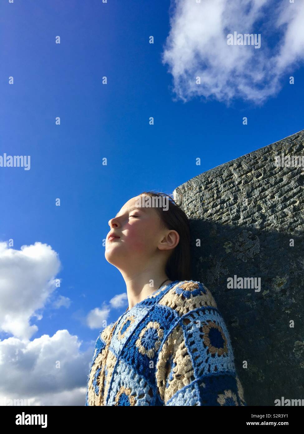 Una joven con los ojos cerrados, apoyada contra una roca plana con el cielo azul en un terreno atrás Foto de stock