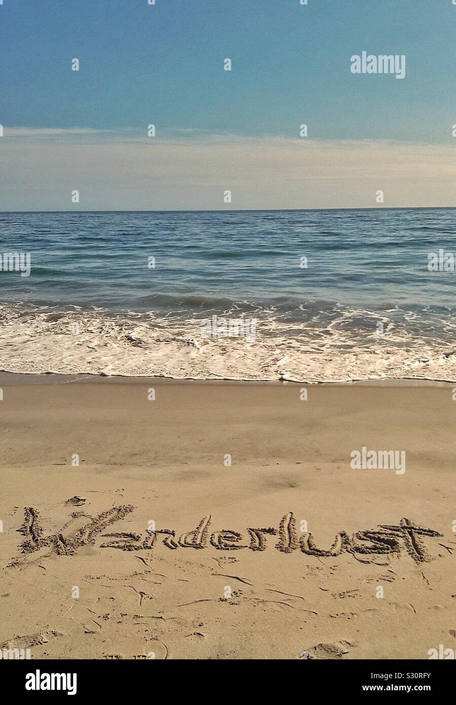 La palabra Wanderlust escrito en la arena de la playa, con vistas al océano. Foto de stock