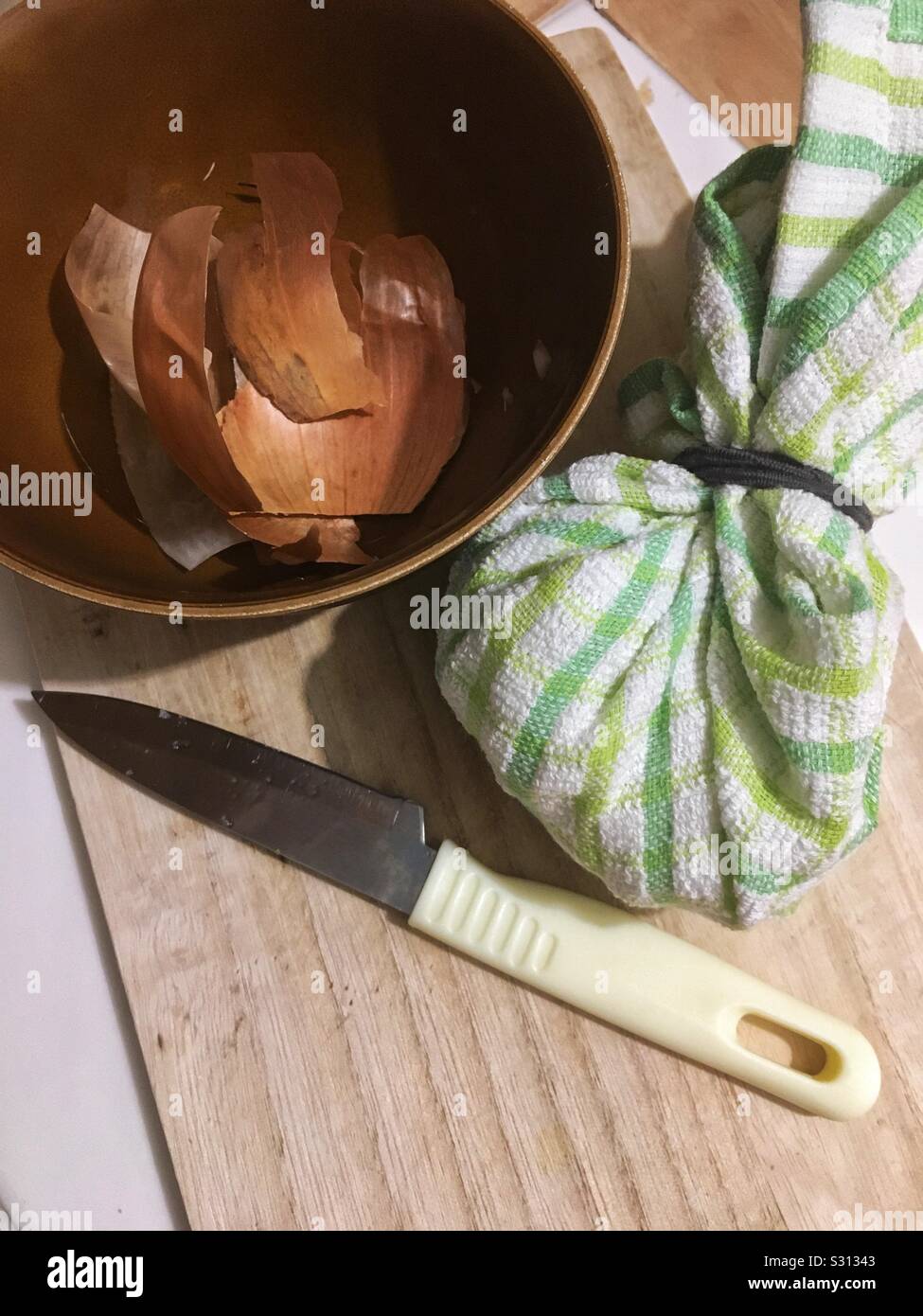 Una tabla de cortar con un cuchillo, una toalla de cocina bundle y un tazón con piel de cebolla, simbolizando la elaboración de un remedio casero para dolor de oído Foto de stock