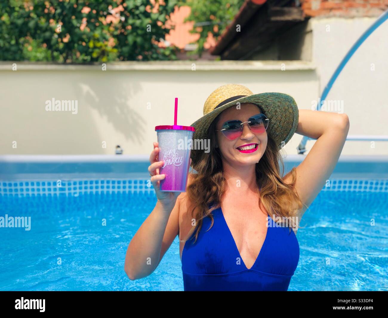 Mujer con bikini azul, gafas de sol y sombrero de paja, sosteniendo una bebida en la piscina Foto de stock