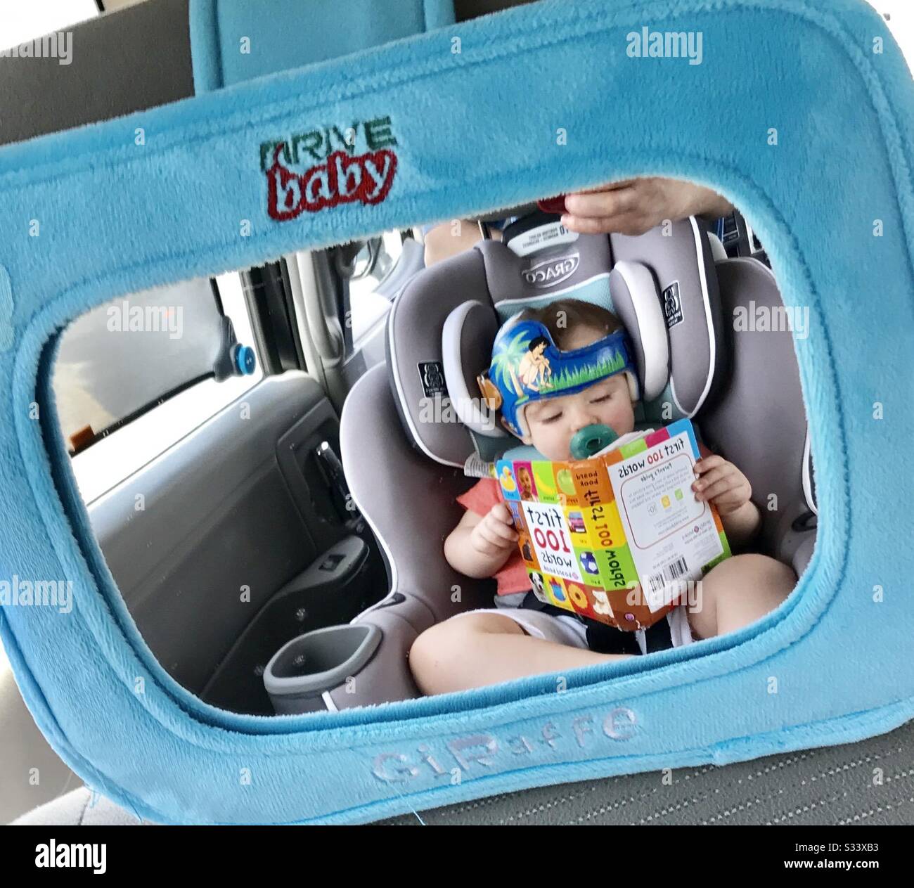 Reflejo del bebé en un coche Murrow mirando el libro. El bebé lleva casco para cabeza plana, plagiocefalia. Foto de stock