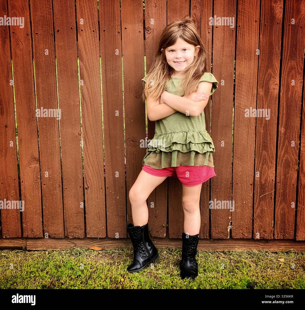 Niña posando su sentido de la moda en un paseo contra una valla de madera marrón manchada. Foto de stock