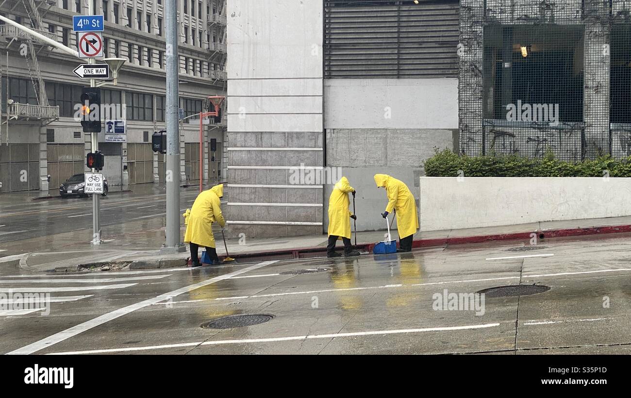 LOS ANGELES, CA, ABR 2020: Tres personas de limpieza en chubasqueros amarillos limpiando canaletas en 4th St, Downtown, al lado de un garaje, bajo la lluvia Foto de stock